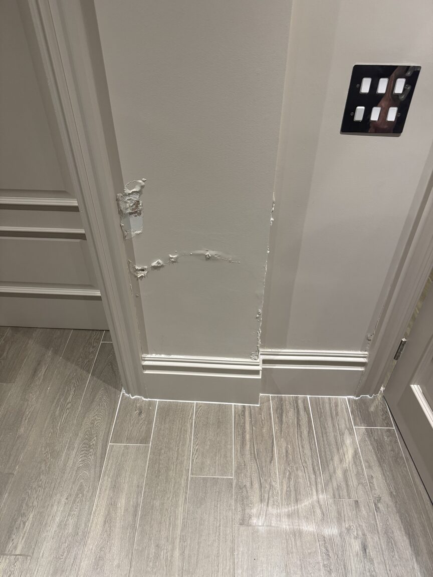 Blown and peeling plaster low on a wall beside a door frame, exposed by moisture damage