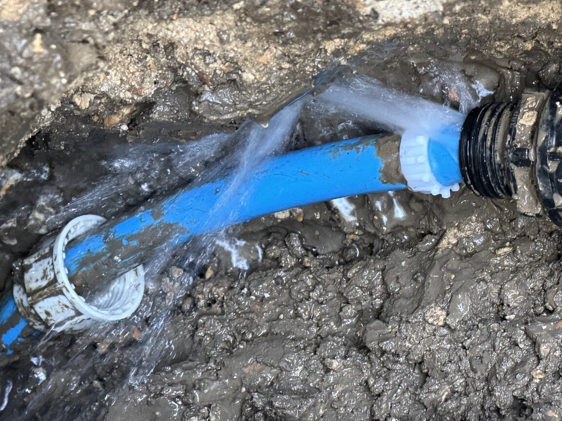 Burst blue plastic water main spraying water inside an open trench during a leak repair