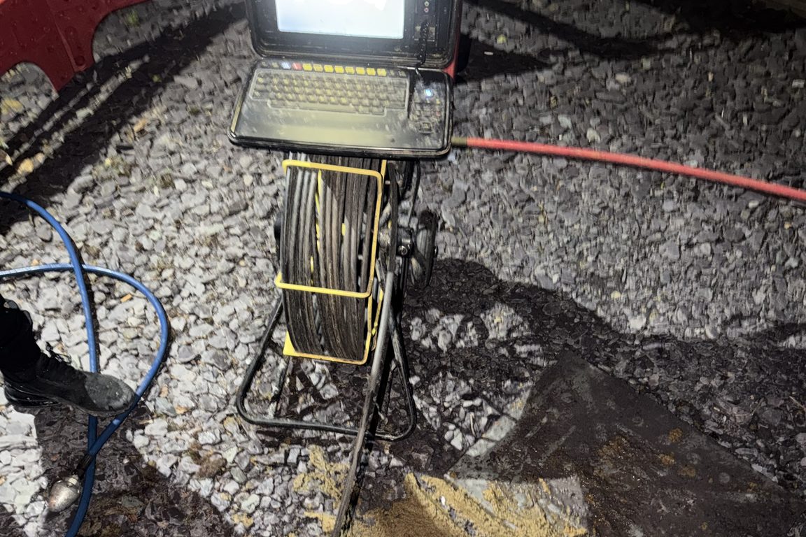 CCTV drain survey camera reel and monitor set up on gravel beside an open chamber at night
