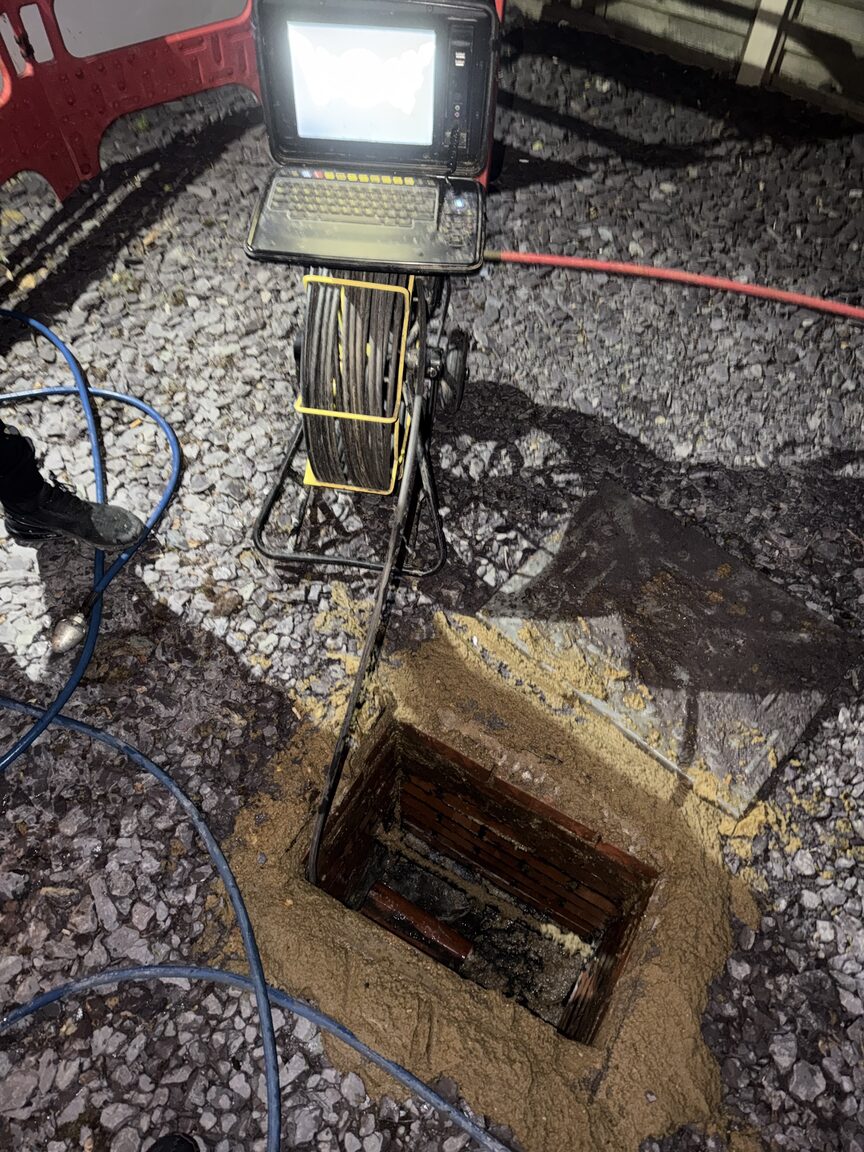 CCTV drain survey camera reel and monitor set up on gravel beside an open chamber at night