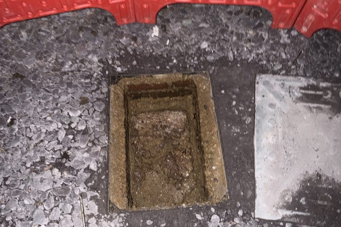 Open drain inspection chamber at night inside red safety barriers, a screwdriver on the gravel beside it