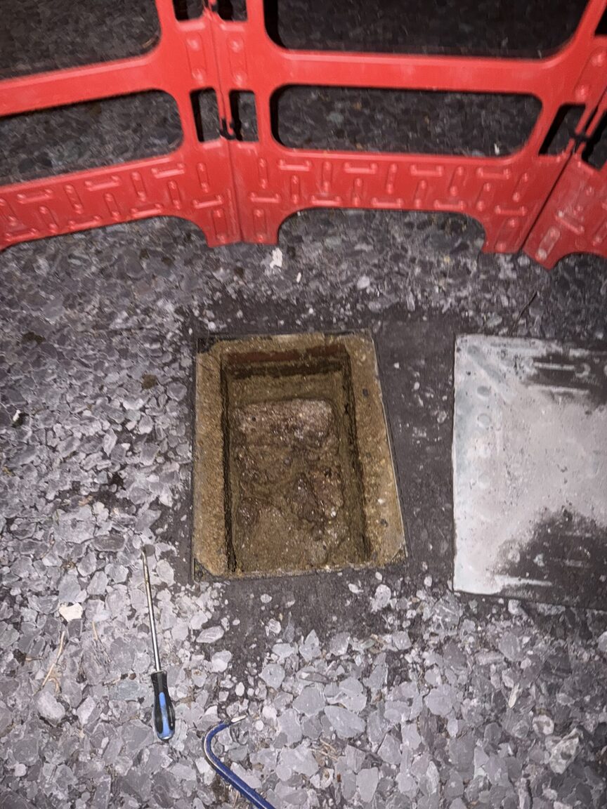 Open drain inspection chamber at night inside red safety barriers, a screwdriver on the gravel beside it