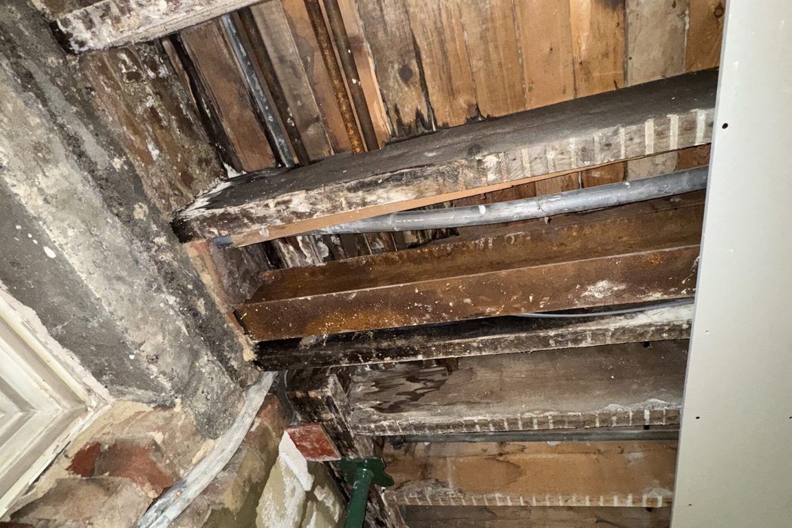 Exposed ceiling joists and steel beams with old water staining, a prop supporting the structure