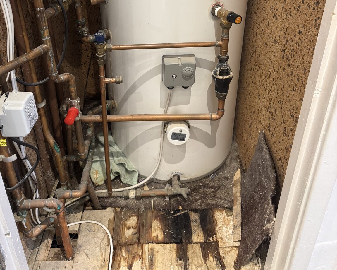 Unvented hot water cylinder with copper pipework and a thermostat, water-stained floorboards and a disturbed fitting beneath it