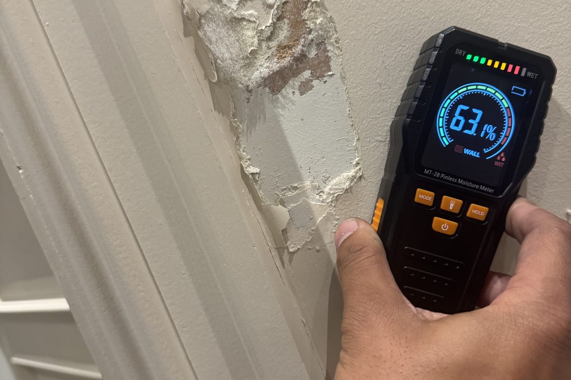 Hand holding a pinless moisture meter reading 63 per cent against blown plaster beside an architrave