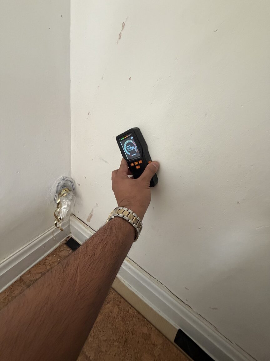 Hand holding a pinless moisture meter reading 60 per cent on a wall just above the skirting board